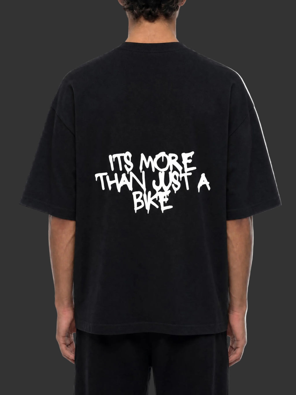 A person shown from the back wearing an oversized black T-shirt with a large white graphic text print reading 'ITS MORE THAN JUST A BIKE' on the back, paired with black pants against a plain white background.