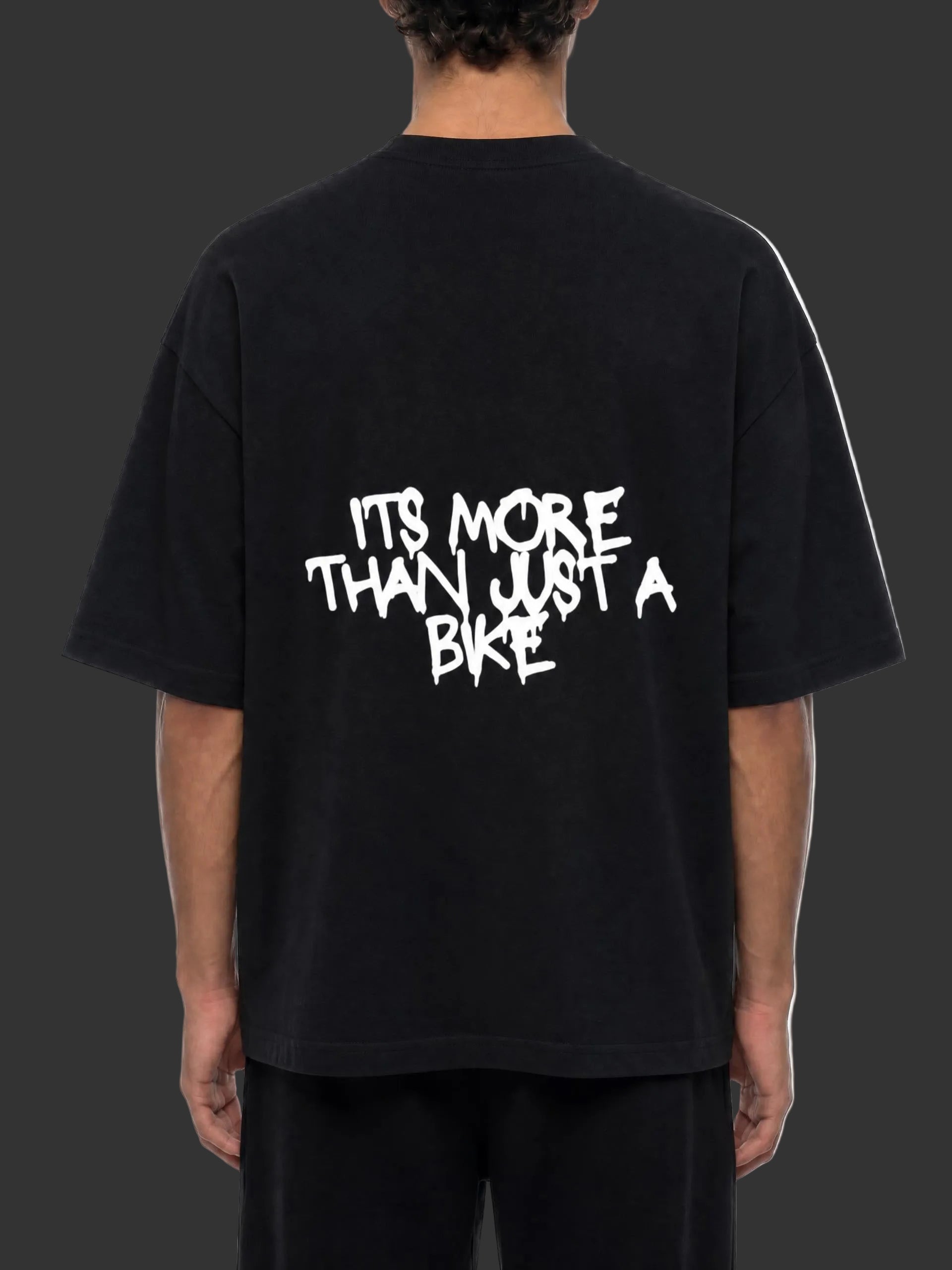 A person shown from the back wearing an oversized black T-shirt with a large white graphic text print reading 'ITS MORE THAN JUST A BIKE' on the back, paired with black pants against a plain white background.